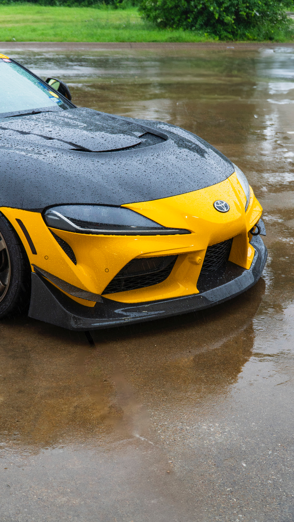 Close-up of a yellow Toyota Supra A90 with a black carbon fiber hood and front splitter, wet from rain.