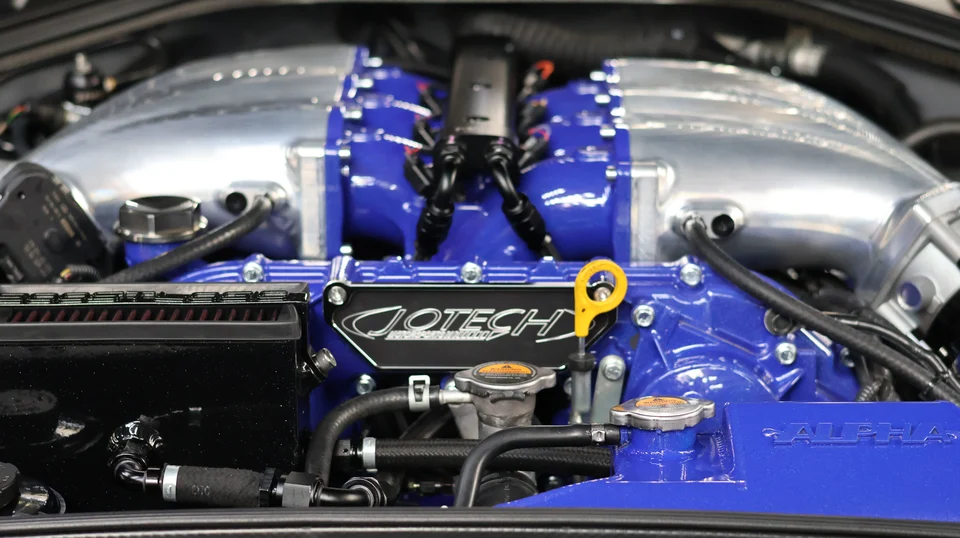 A close-up of a highly modified Nissan GT-R engine bay featuring a blue Jotech Motorsports intake manifold and Alpha Performance components.