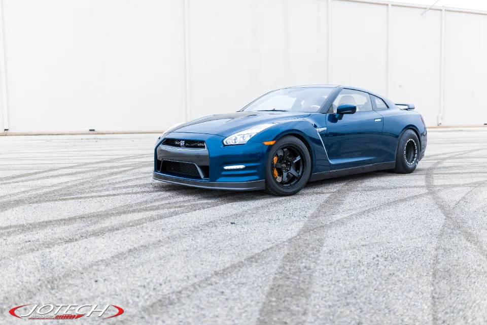 A blue Nissan GT-R with black drag wheels and a front splitter parked on an asphalt lot with tire marks against a white wall.