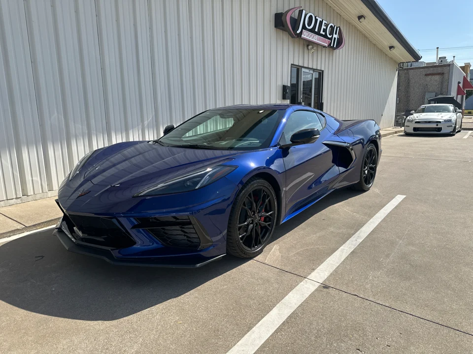 A blue Chevrolet Corvette C8 parked outside Jotech Motorsports, with a white Nissan GT-R visible in the background.