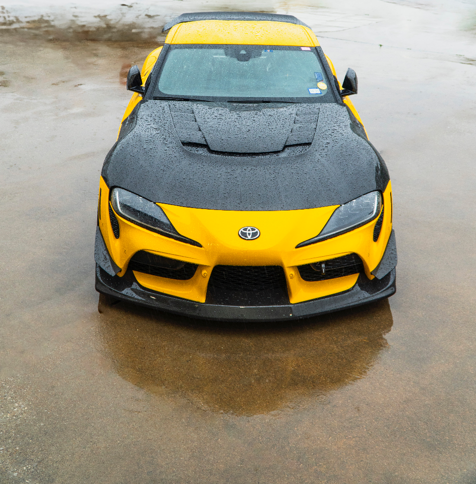 Front view of a yellow Toyota Supra MK5 with a black carbon fiber hood and front splitter parked on wet pavement.