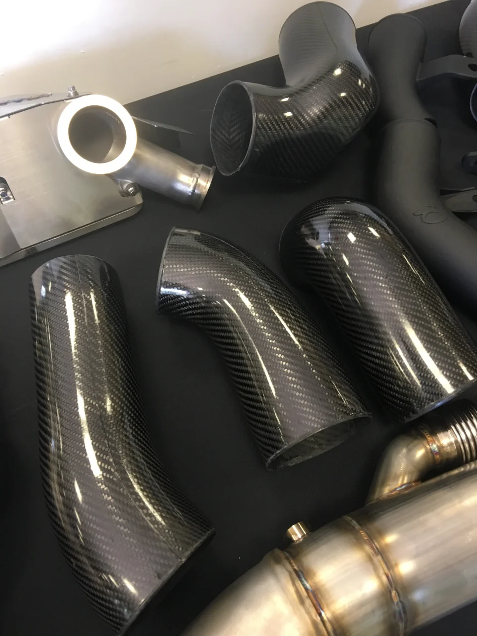 Close-up of various high-performance car parts including carbon fiber intake pipes and stainless steel exhaust components laid out on a dark surface.