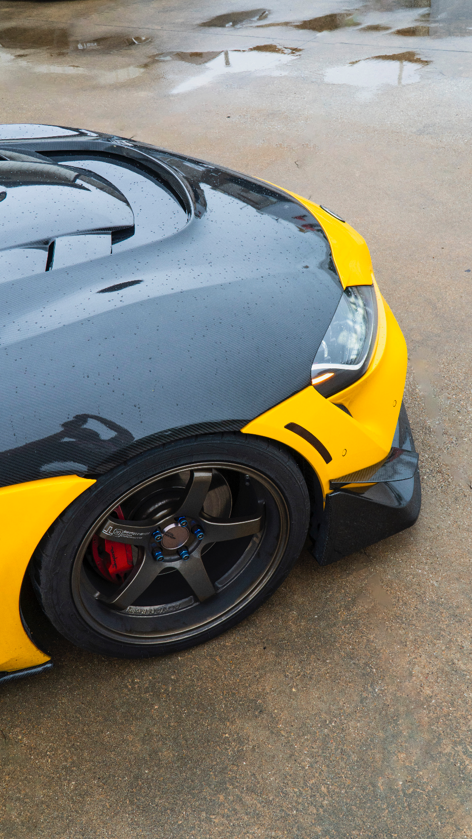 Close-up of a custom car featuring a carbon fiber hood, yellow fender, aftermarket wheel, and red brake calipers.