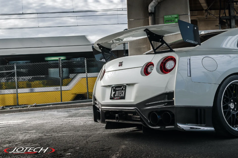 Rear view of a white Jotech Motorsports Nissan GT-R with a large carbon fiber wing and aftermarket exhaust, parked near train tracks.
