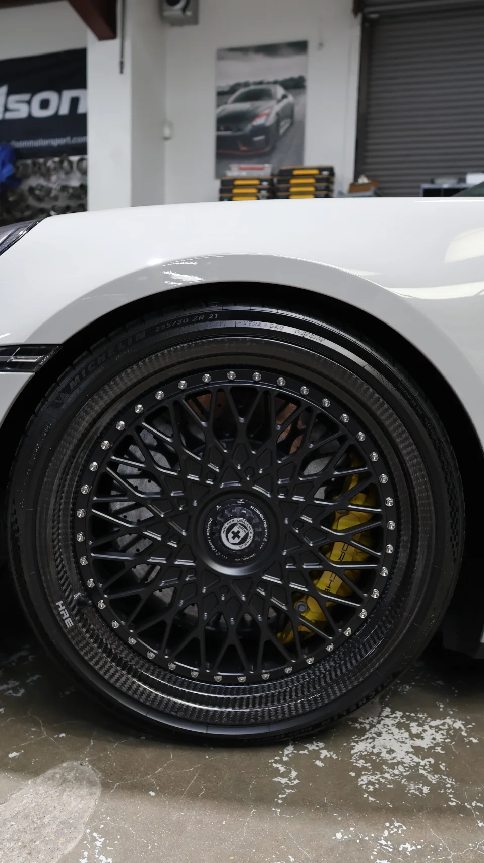 A white Porsche with HRE carbon fiber wheels and yellow brake calipers in a shop setting, showcasing the wheel detail.