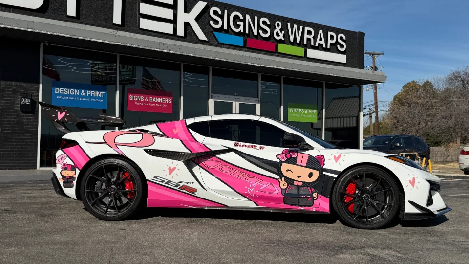 A white and pink custom-wrapped Corvette C8 with a large rear wing parked in front of a Jotech Signs & Wraps shop.