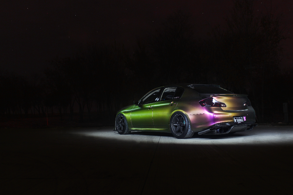Infiniti G37 sedan with a color-shifting chameleon wrap, custom wheels, and a rear diffuser, parked at night.