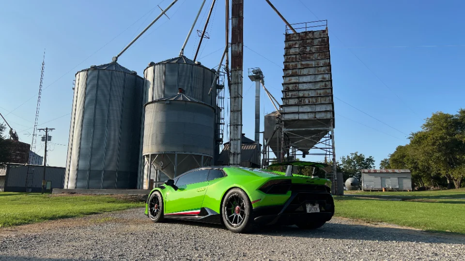 A vibrant green Lamborghini Huracan with an aftermarket wing and custom wheels parked on a gravel road in front of large grain silos.