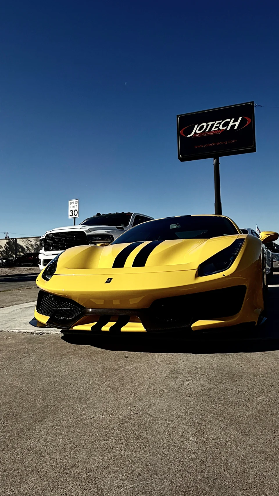 A bright yellow Ferrari sports car with black racing stripes parked outdoors in front of a Jotech Motorsports sign.