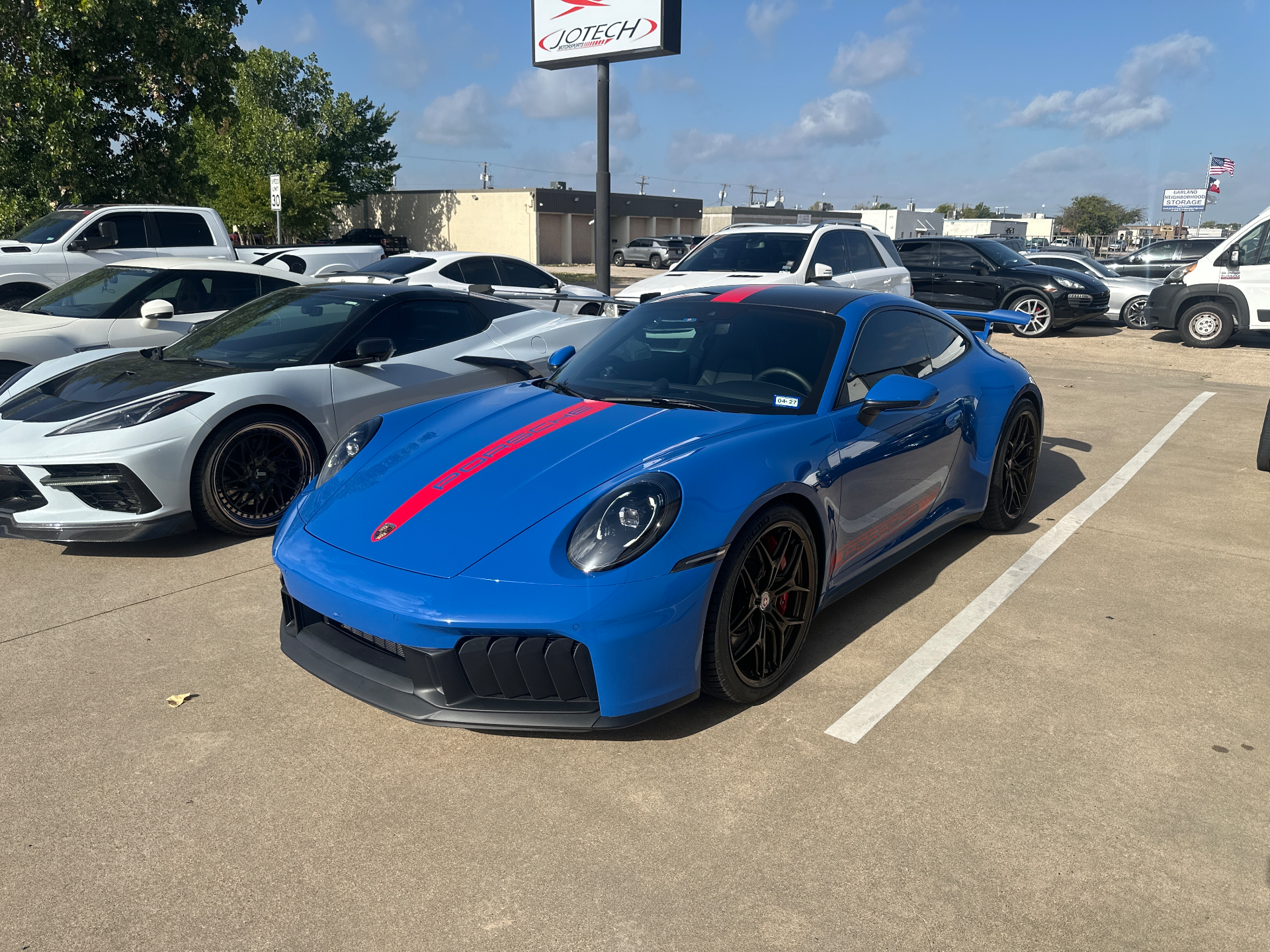 A blue Porsche 911 GT3 RS with red racing stripes parked in a lot outside Jotech Motorsports, with a white C8 Corvette nearby.