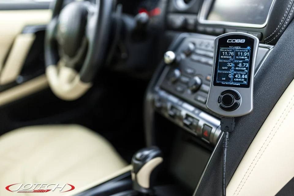A Cobb Accessport tuning device displaying live data, plugged into the dashboard of a Nissan GT-R interior.