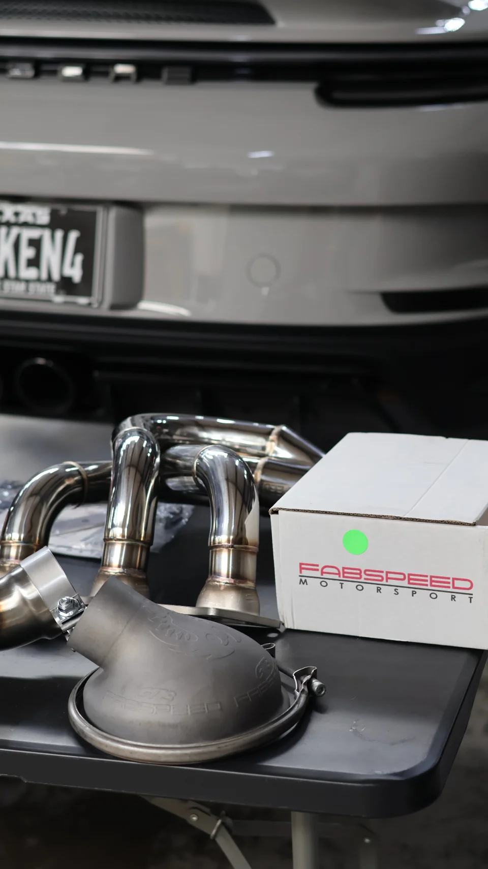 Fabspeed Motorsport exhaust headers and performance parts on a table in front of a grey Porsche, ready for installation.