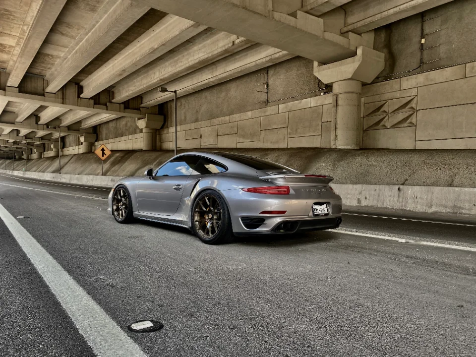 Silver Porsche 911 Turbo S with aftermarket wheels parked under a concrete overpass.