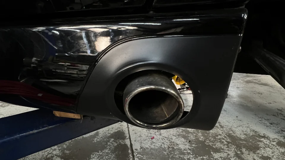 A close-up detail shot of a large, chrome exhaust tip integrated into the matte black rear bumper of a black car, likely in a shop.