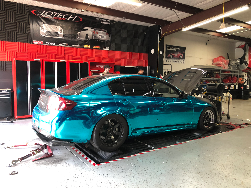 A teal chrome wrapped Infiniti G37 sedan on a dyno at Jotech Motorsports for performance tuning, with the hood open.