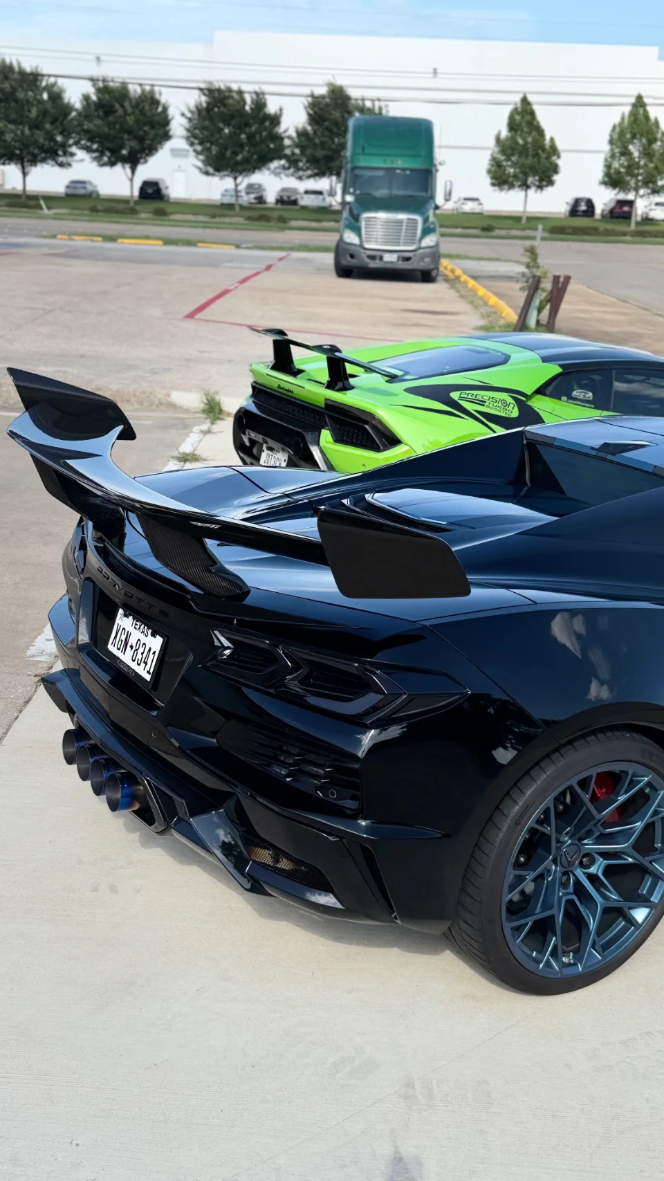 A black C8 Corvette with a large carbon fiber wing and custom blue wheels parked next to a green Lamborghini Huracan.