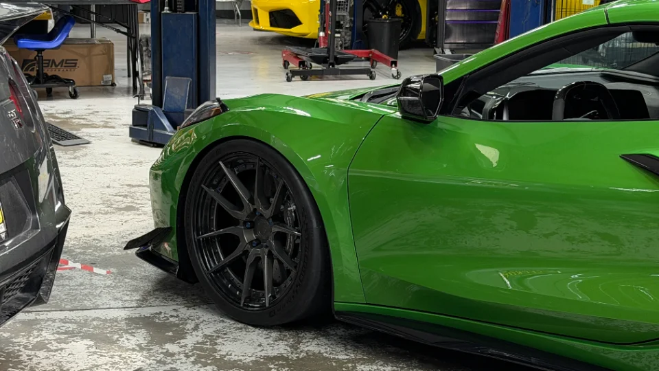 A green C8 Corvette with aftermarket wheels and a front splitter parked in a performance tuning shop.