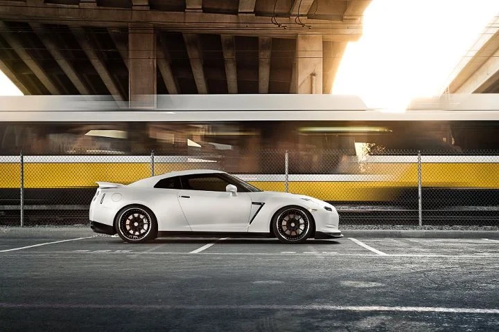 White Nissan GT-R parked in an urban setting with a blurred train passing by in the background.