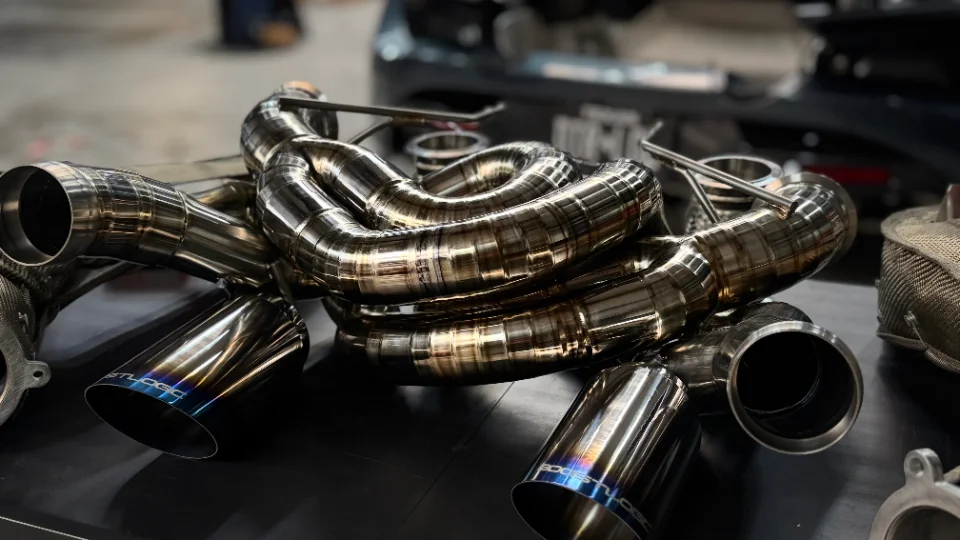 A close-up of a polished titanium aftermarket exhaust system with blue-tinted tips, likely for a high-performance car.