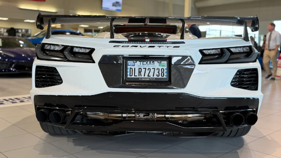 Rear view of a white C8 Corvette with a large carbon fiber wing and aftermarket exhaust in a showroom.