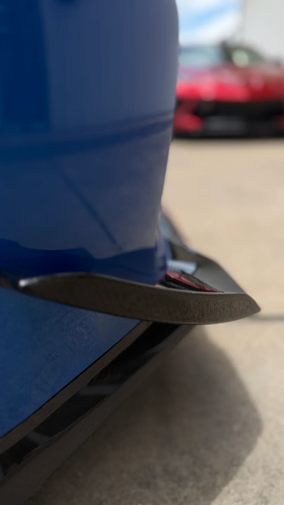 A close-up detail shot of a blue car's rear with a carbon fiber diffuser, with a red car blurred in the background.