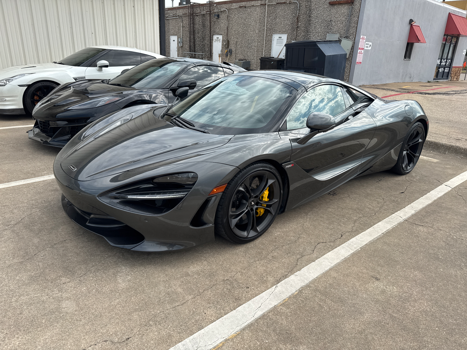 A grey McLaren 720S, black C8 Corvette, and white Nissan GT-R parked in a lot outside a building.