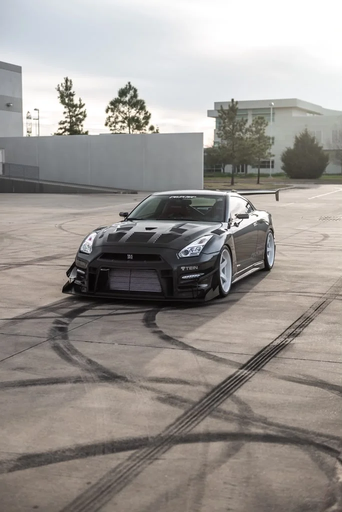 A black widebody Nissan GT-R with aftermarket wheels and custom aero parked in a large lot with tire marks.