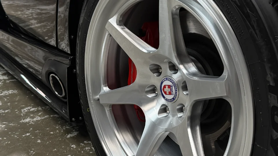 Close-up of a black car's HRE performance wheels with red brake calipers and a carbon fiber side skirt.