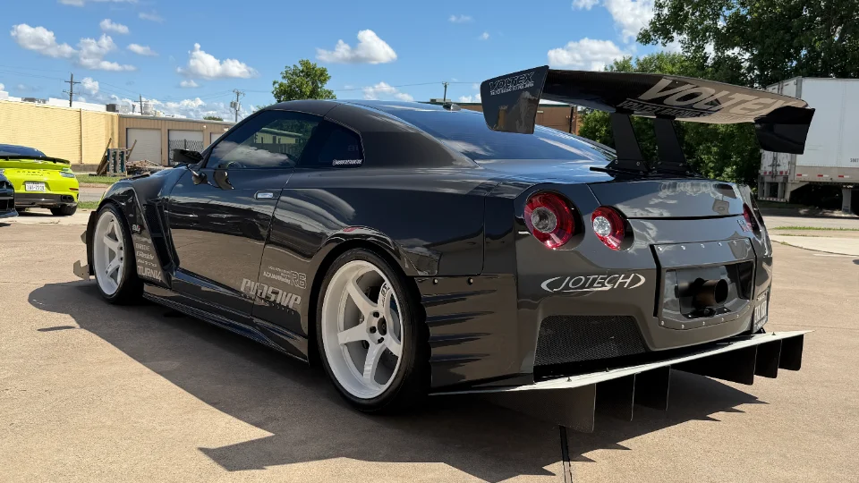 A dark grey widebody Nissan GT-R with a large Voltex carbon fiber wing, aggressive rear diffuser, and white aftermarket wheels.