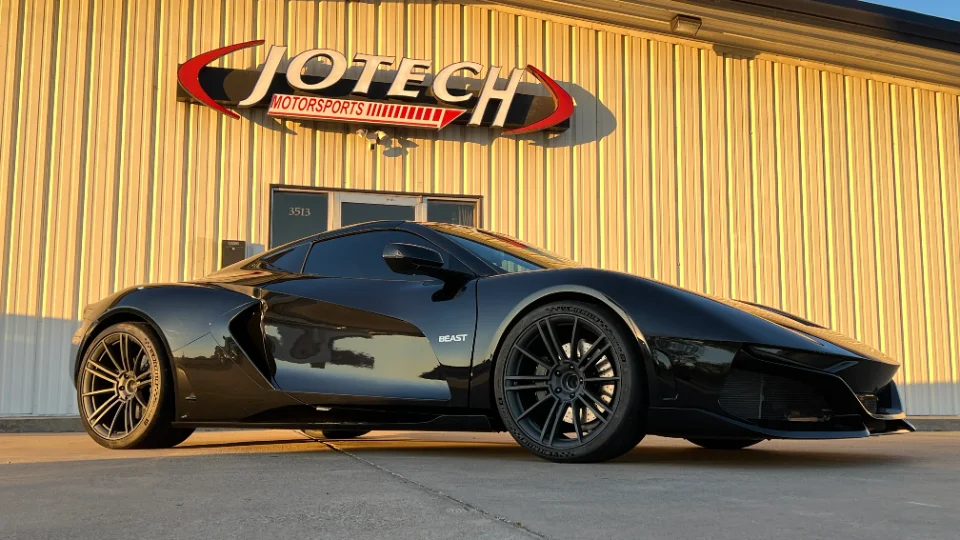 A sleek black exotic sports car, possibly a custom build, parked in front of the Jotech Motorsports facility at sunset.