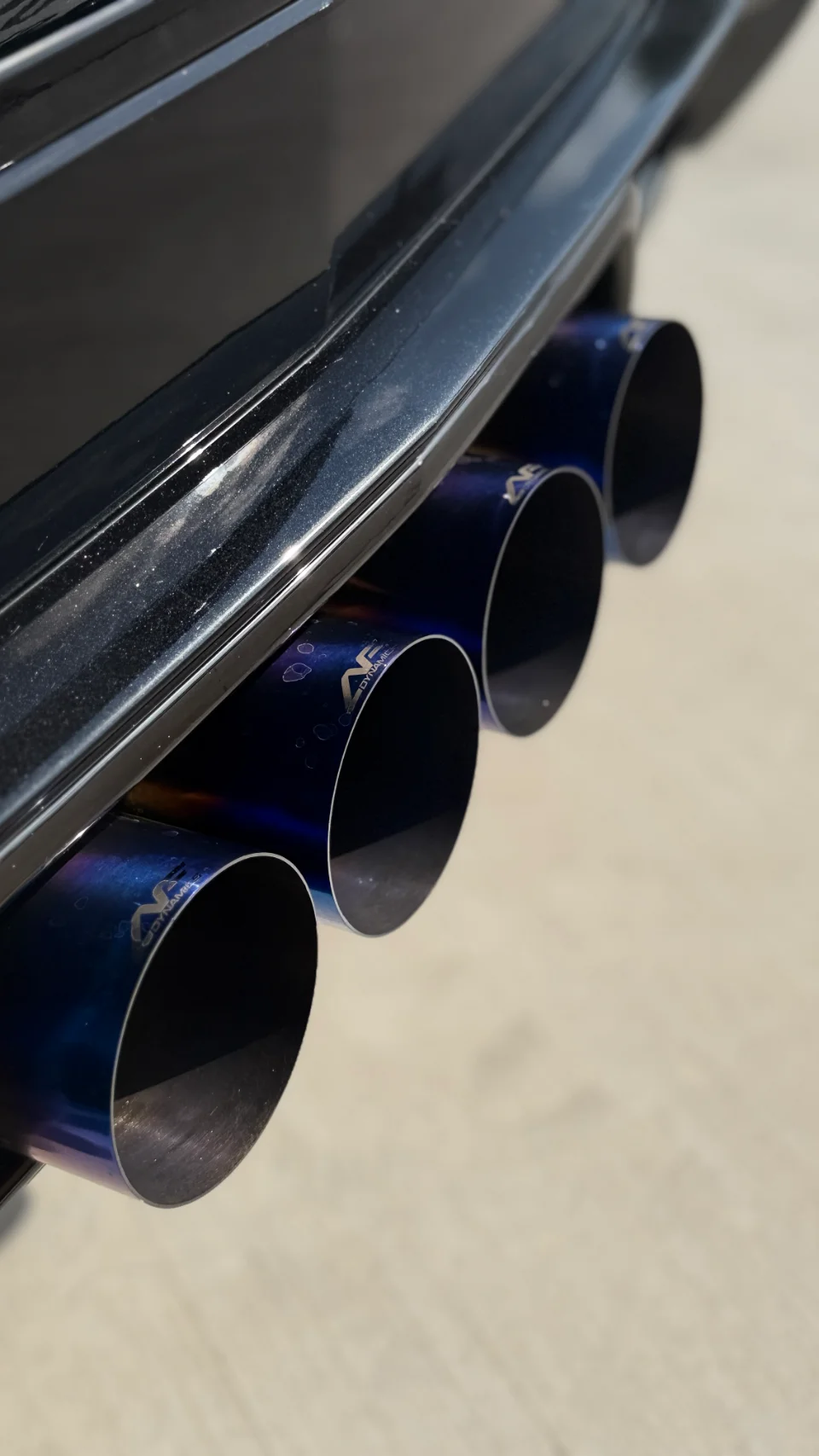 Close-up of AWE Tuning quad titanium exhaust tips on a performance car, showing the blue burnt finish.