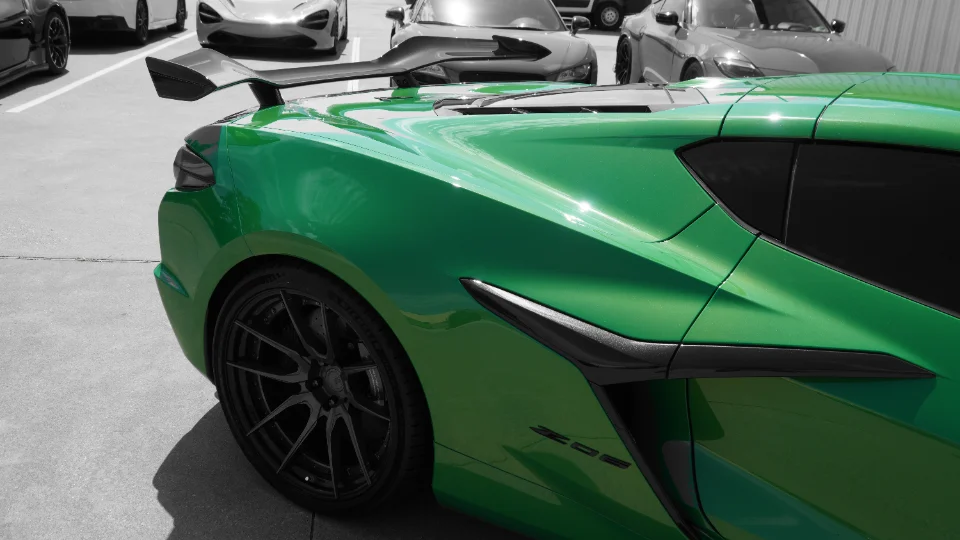 A vibrant green Corvette C8 Z06 with a large rear wing and black multi-spoke wheels, parked outdoors with other luxury cars.