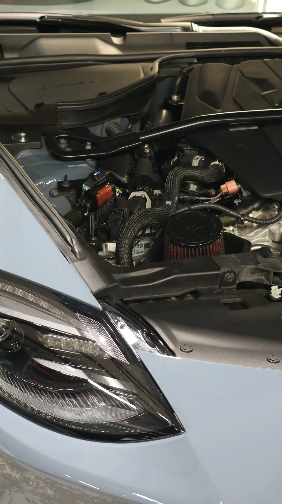 A close-up view of a light blue car's engine bay, showcasing an aftermarket air intake system with a red filter.