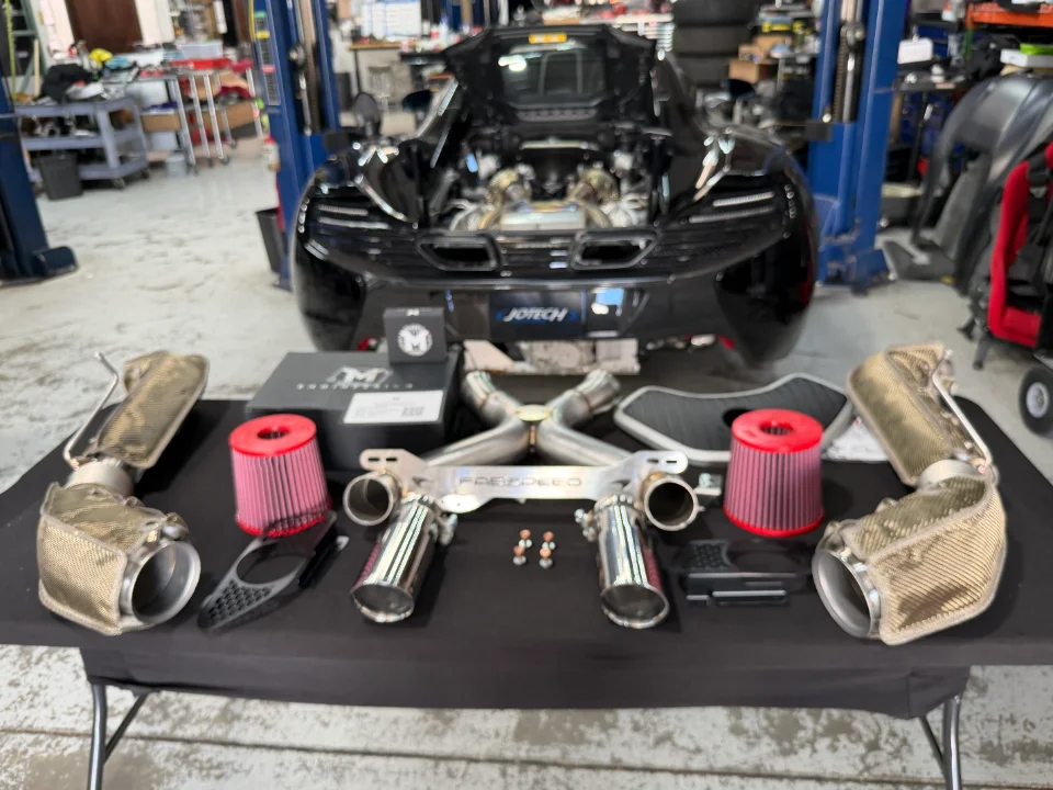 A black McLaren sports car on a lift in a Jotech Motorsports shop, with an aftermarket exhaust system and air filters on a table.
