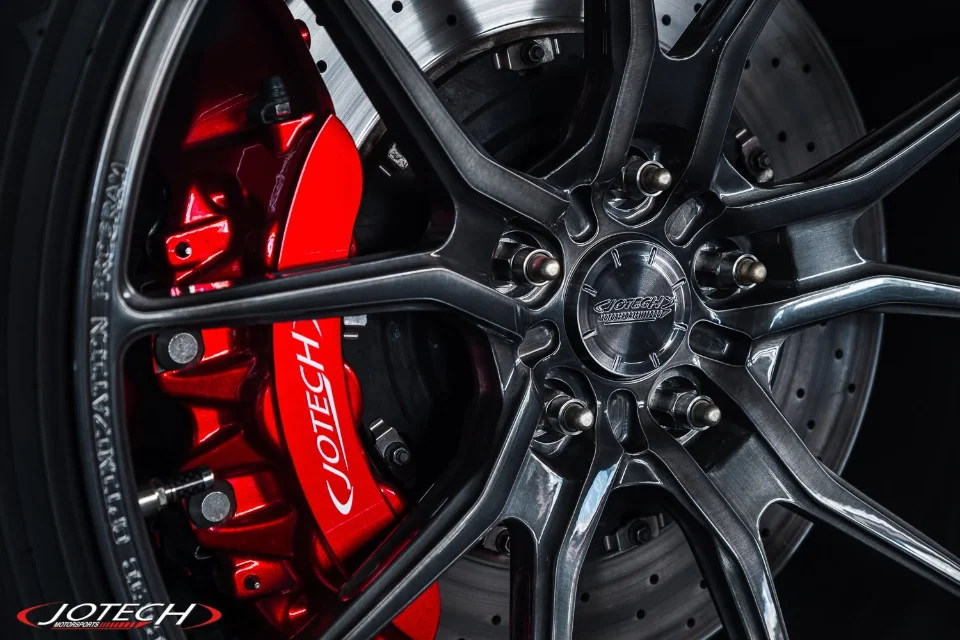 Close-up of a car wheel with dark spokes and a bright red Jotech brake caliper and drilled rotor.