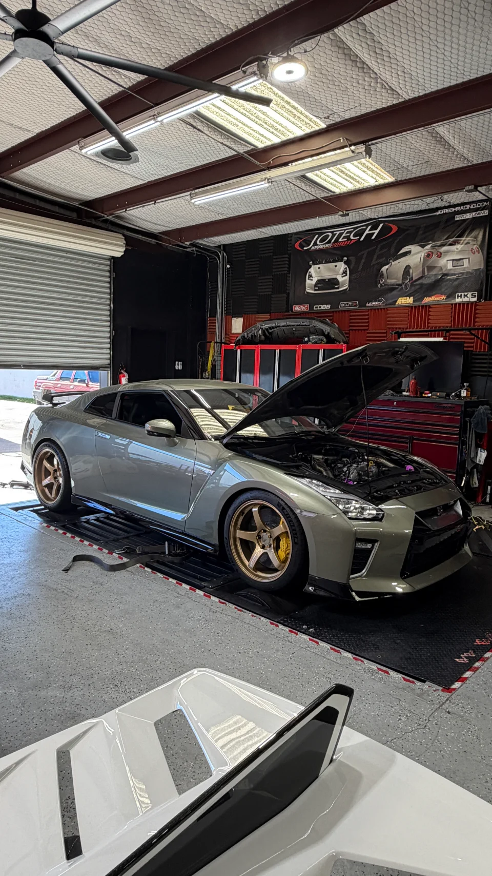 A grey Nissan GT-R with its hood open, strapped to a dyno in the Jotech Motorsports shop.