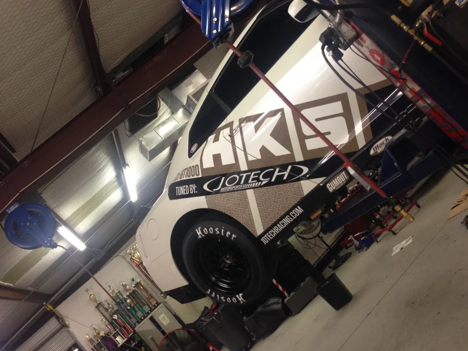 A white Jotech Motorsports race car with HKS livery and Hoosier drag tires on stands in a shop with trophies in the background.
