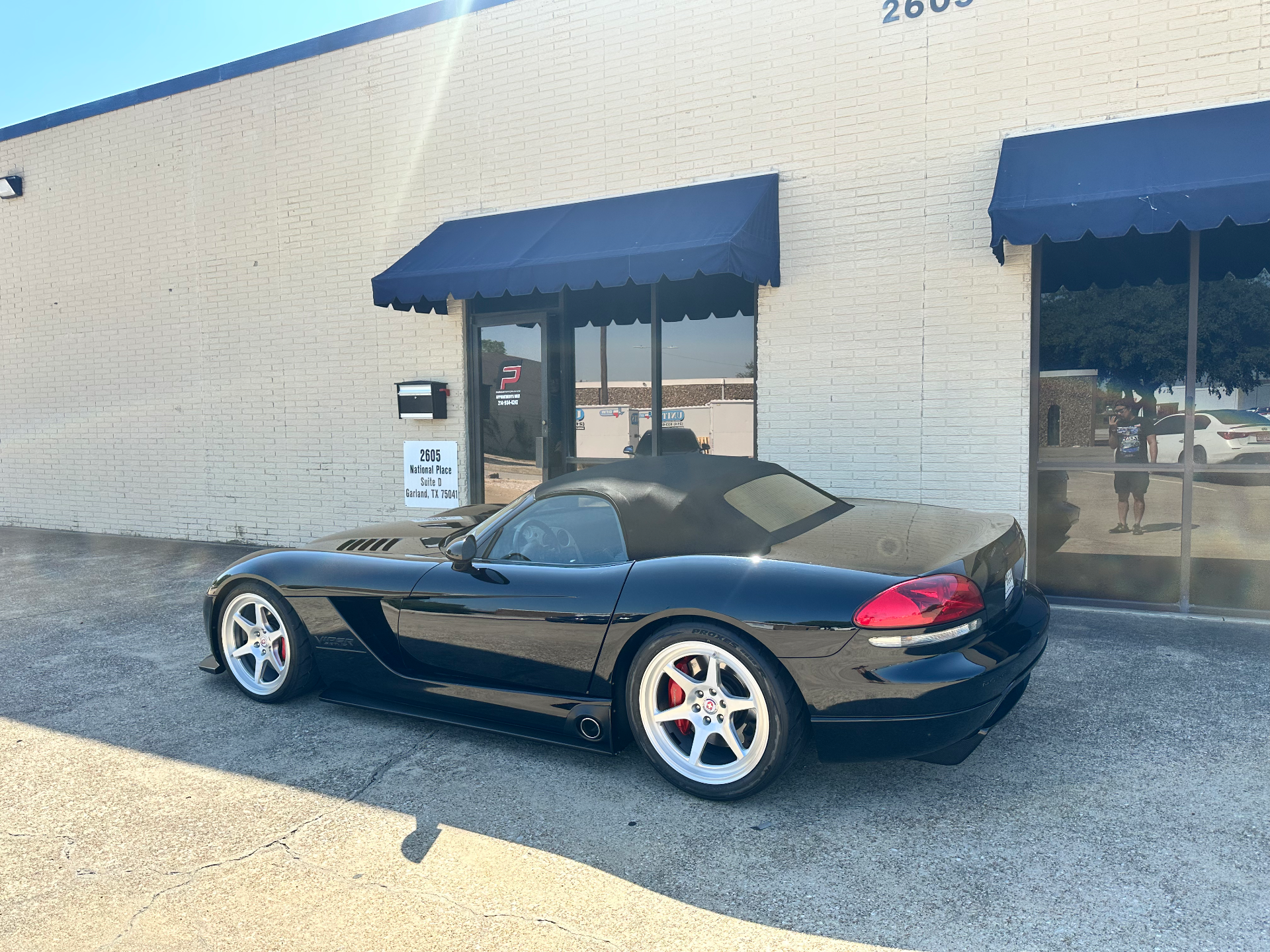 A black Dodge Viper convertible with aftermarket wheels parked outside a performance shop on a sunny day.