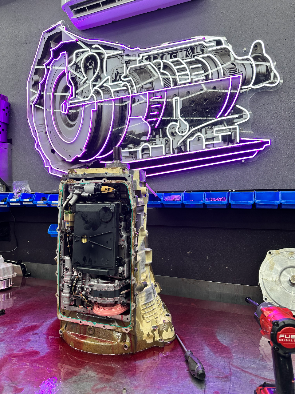 An open car transmission on a workbench in a performance tuning shop, with a neon diagram on the wall.