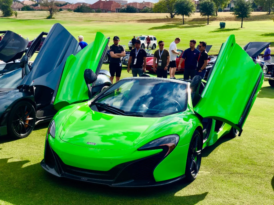 Bright green McLaren supercar with its unique doors open, displayed on a grassy field at an outdoor car show event with people around.