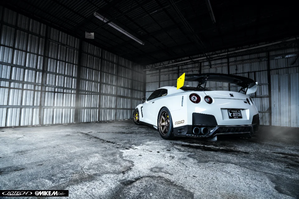 A white Nissan GT-R with a large rear wing and aftermarket wheels parked in an industrial warehouse.