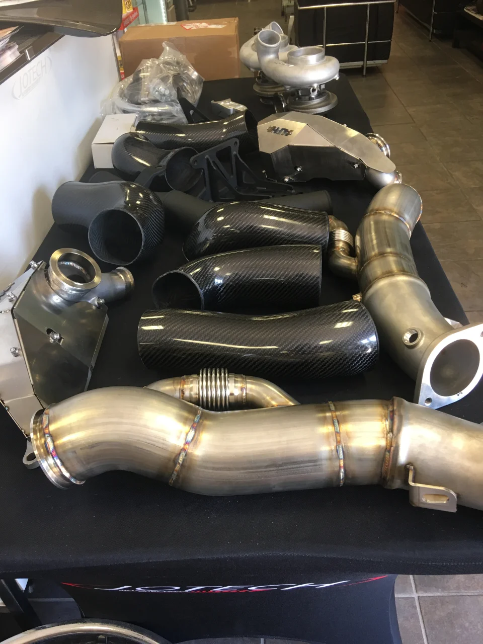 A collection of high-performance car parts including carbon fiber intake pipes, turbochargers, and stainless steel exhaust downpipes on a table.