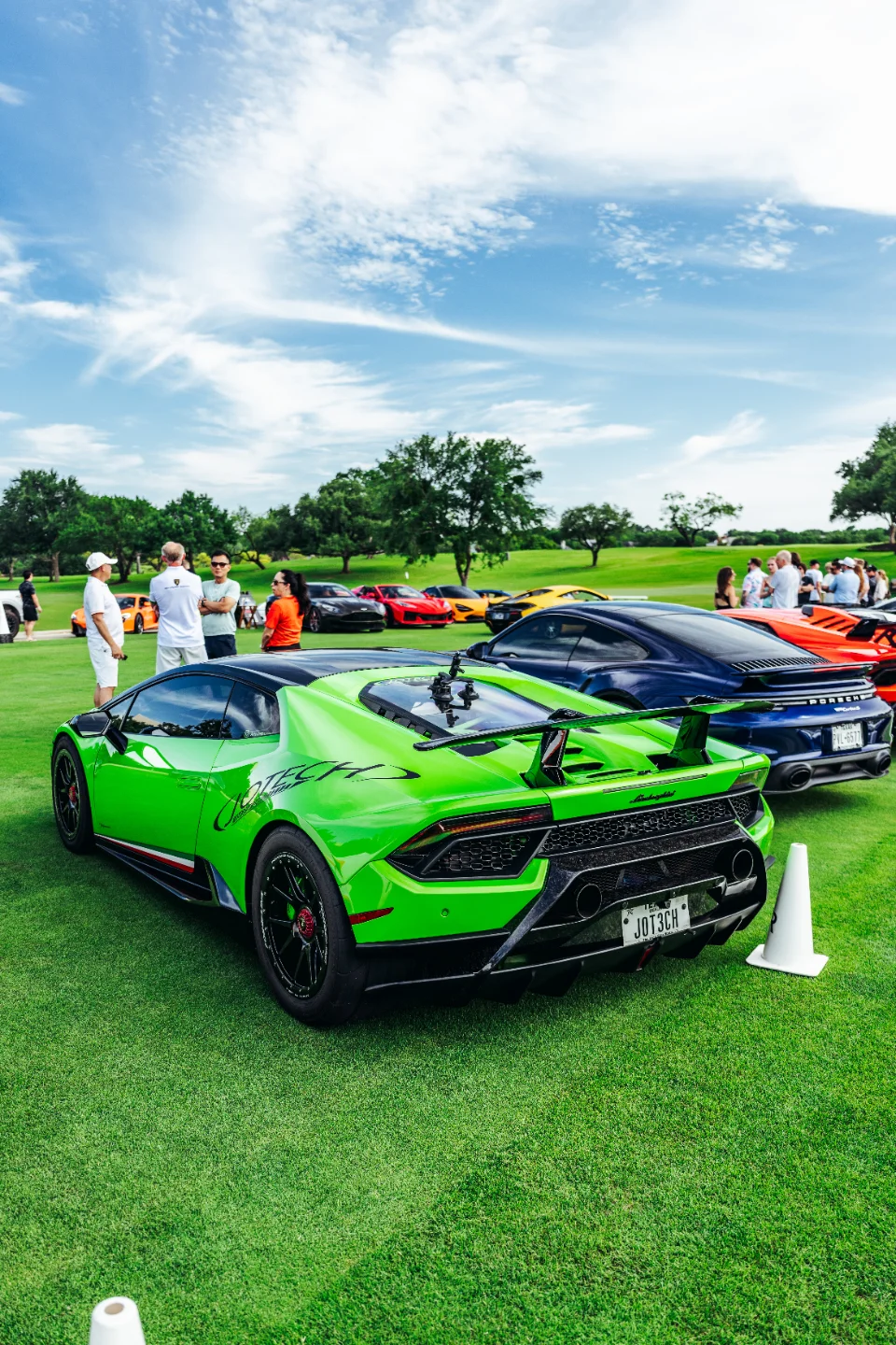 A green Lamborghini Huracan with a large rear wing parked on grass at an outdoor car show with other luxury cars and people.
