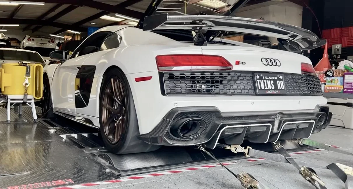 A white Audi R8 supercar with a large carbon fiber wing strapped down on a dynamometer for performance testing.