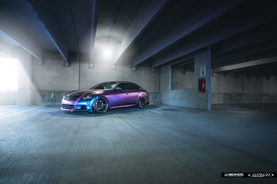 A custom Infiniti G37 sedan with a vibrant color-shifting wrap and aftermarket wheels parked in a concrete garage.