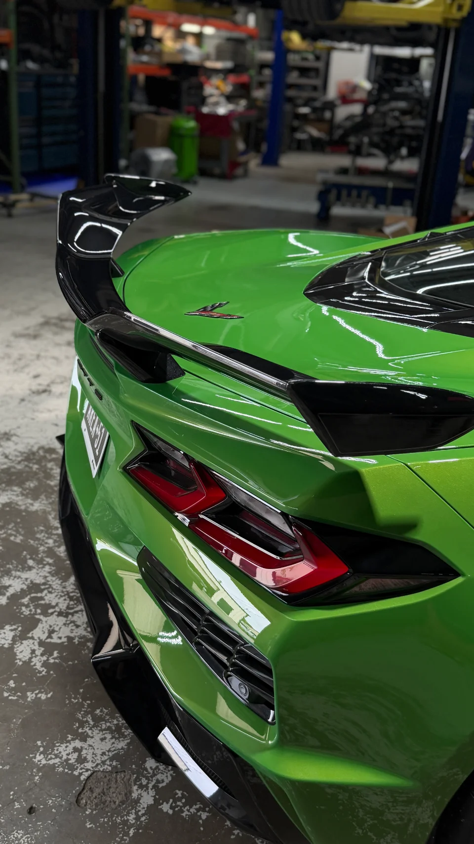 Rear view of a vibrant green C8 Corvette with a large carbon fiber wing, parked inside a performance shop.