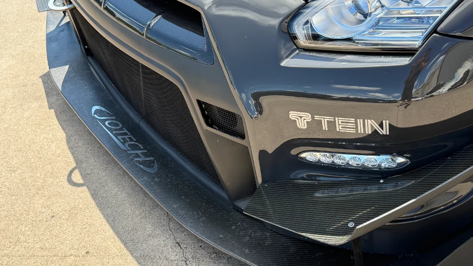 Close-up of a black Nissan GT-R front bumper with carbon fiber splitter, Jotech branding, and Tein logo.
