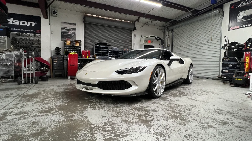 White Ferrari 296 GTB parked inside a performance tuning shop with garage doors and equipment in the background.