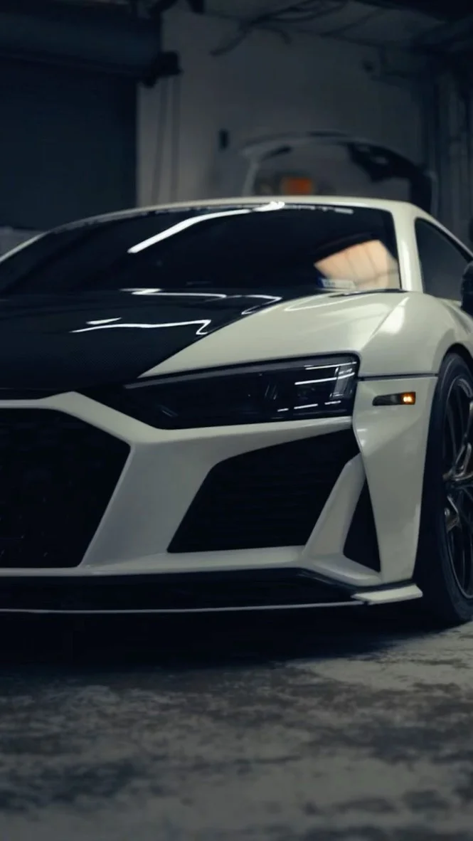 Front view of a white Audi R8 with a black carbon fiber hood and aggressive front bumper in a garage setting.