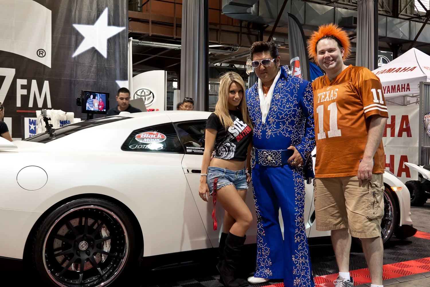 People posing with a white Nissan GT-R at an event, including an Elvis impersonator and a woman in denim shorts.